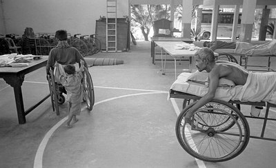 Un homme handicapé en fauteuil roulant est porté par son fils; un autre homme dans la même situation le regarde au Centre de Réhabilitation des Paralysés, CRP à Savar, Dhaka, Bangladesh. 2005. - Unbekannt Unbekannt